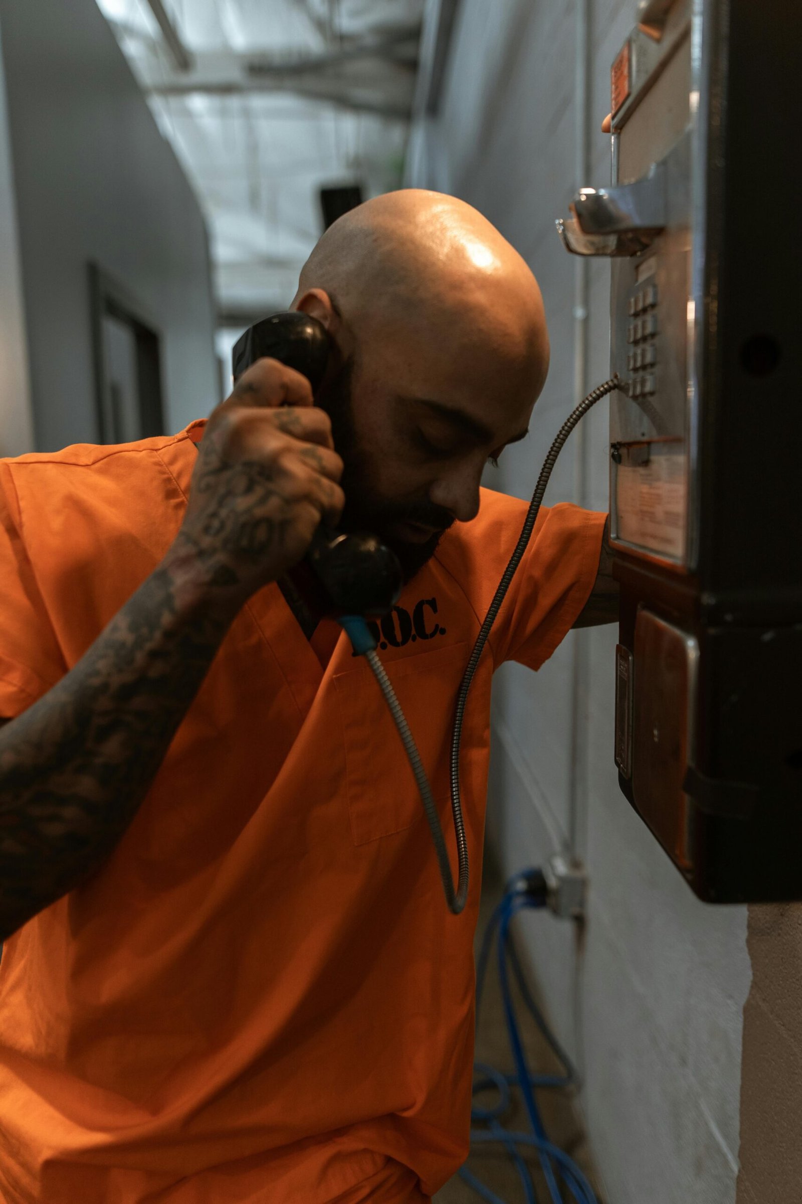 A tattooed male prisoner in orange uniform uses a telephone inside a jail facility.
